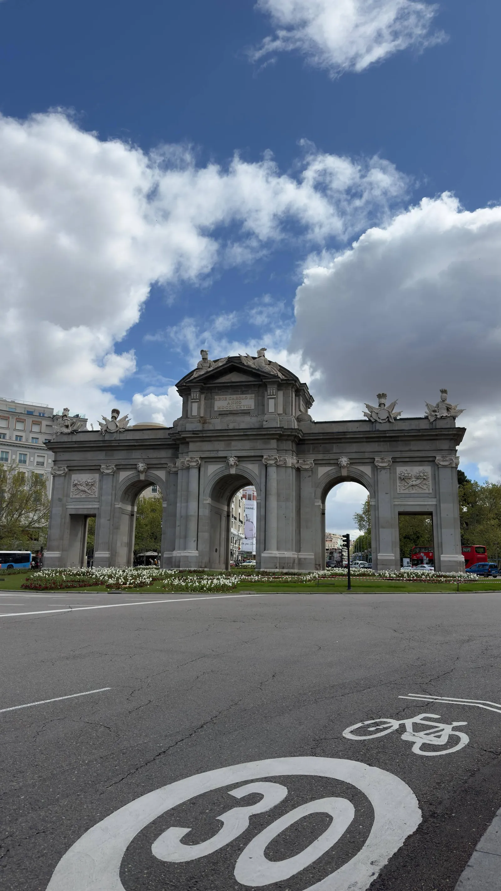 Puerta de Alcalá bajo un cielo azul con nubes, flores en primer plano