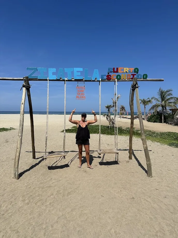 Dani posando bajo el cartel de Zicatela Puerto Escondido en la playa con palmeras al fondo